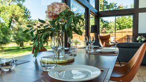 Une table à manger dressée avec des assiettes, des verres et un vase de fleurs, donnant sur des arbres et une clôture en bois à l’extérieur.