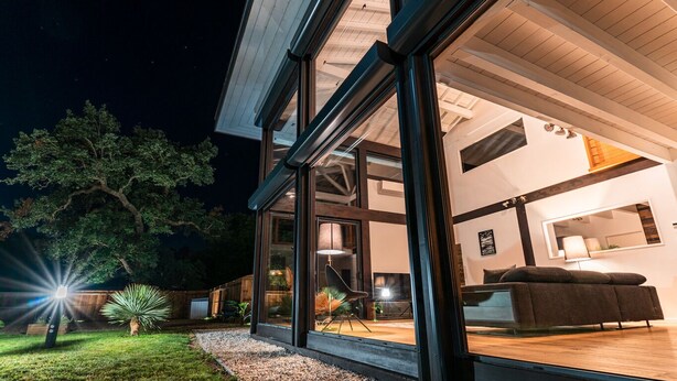 Une maison moderne dotée de grandes baies vitrées, d’une terrasse en bois et d’un intérieur bien éclairé.