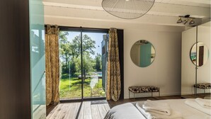 Une chambre avec un grand lit, un miroir rond et une vue sur un jardin, accessible par une porte-fenêtre coulissante.