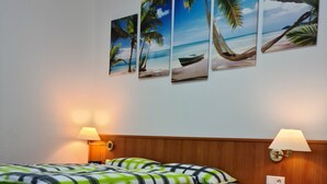 A neatly made bed with a patterned bedspread, two bedside lamps, and a wall with four framed tropical beach scenes. - Pension Kreihe (Bad Lauterberg)