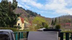 A balcony with wicker furniture and a glass table, overlooking a mountainous landscape with buildings. - Pension Kreihe (Bad Lauterberg)