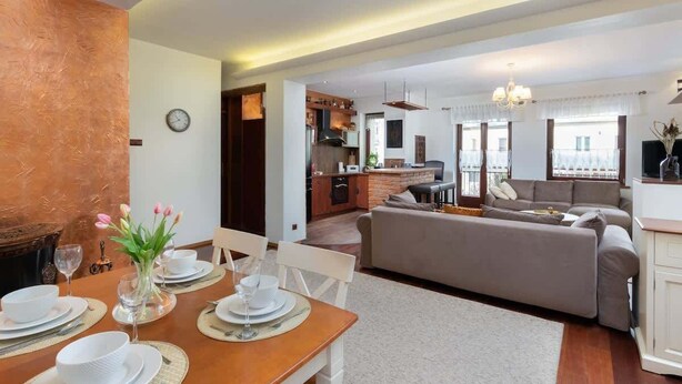 A modern living room with a sofa, dining table set for four, and a kitchen in the background. - Comfortable Apartment in Gdynia | 2 Bedrooms | Parking (Gdynia)