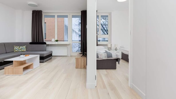 Ein modernes Wohnzimmer mit einem grauen Sofa, einem weißen Couchtisch und einem Holznachttisch. Ein Fenster bietet Blick auf ein Backsteinhaus und Bäume.
