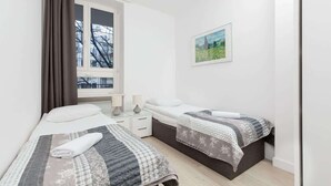 Ein Schlafzimmer mit zwei Betten, einem Fenster mit Vorhängen, einem Nachttisch mit Lampen und einem gerahmten Bild an der Wand.