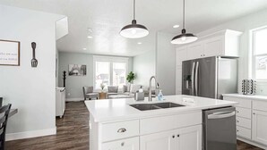 Una cocina moderna con armarios blancos, una isla central con lavamanos, electrodomésticos de acero inoxidable, iluminación colgante y vistas a una sala de estar.