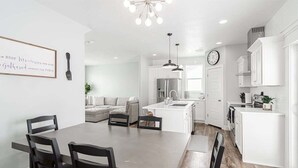 Una cocina moderna con armarios blancos, un comedor con una mesa grande y sillas, y una sala de estar con un sofá y una mesita de centro.