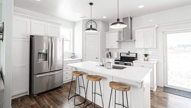 Una cocina moderna con armarios blancos, electrodom ésticos de acero inoxidable, una isla central con taburetes, iluminación colgante y un reloj en la pared.