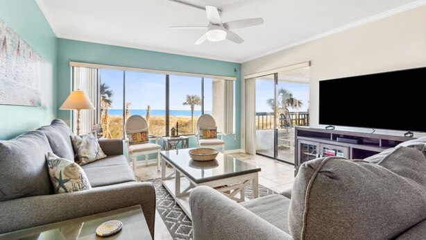 Un vaste espace de vie comprenant un canapé, des fauteuils, une table basse et un téléviseur. Une grande fenêtre offre une vue sur la plage et les palmiers.