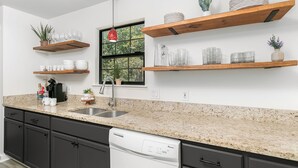A modern kitchen with granite countertops, dark cabinets, and a dishwasher. There are wooden shelves above the counter holding glassware and plants.