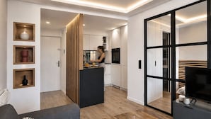 A modern living space with a sofa, coffee table, and a TV. There is a kitchen area with a person preparing food. - VACATELLA - LES PINS ENSOLEILLÉS / Contemporary apartment with sea view (Sainte-Maxime)