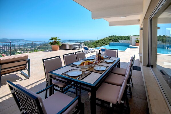 A terrace with a dining table set for four, overlooking a swimming pool and a scenic view. - Luxury Villa Olive with heated infinity pool and Jacuzzi - near Dubrovnik (Ivanica - Ravno)