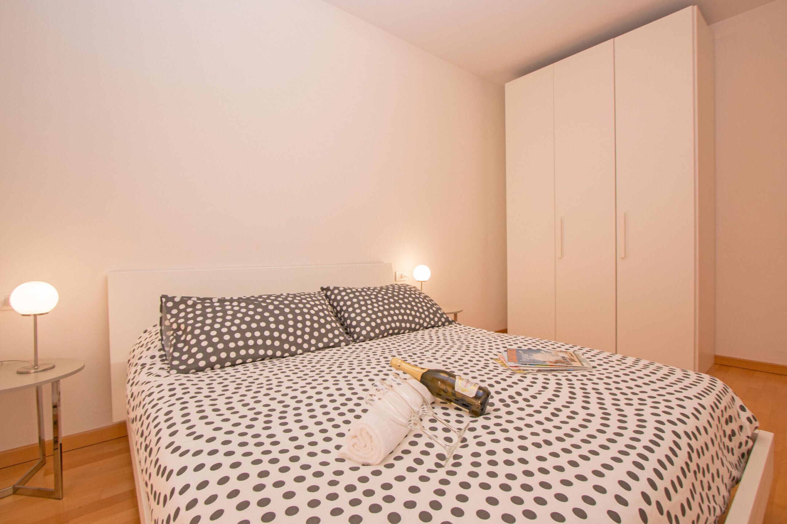 A bedroom with a bed, a nightstand, a lamp, a wardrobe, and a bottle of champagne.
