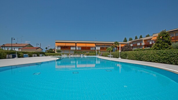 Ein Schwimmbecken mit klarer, blauer Wasseroberfläche, umgeben von einer begrünten Anlage mit Hecken und einem Gebäude im Hintergrund mit orangefarbenen und weißen Außenwänden.