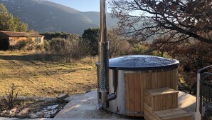 Un bain à remous doté d’une structure extérieure en bois et de tuyaux en métal, installé dans un jardin avec une vue sur les montagnes en arrière-plan.