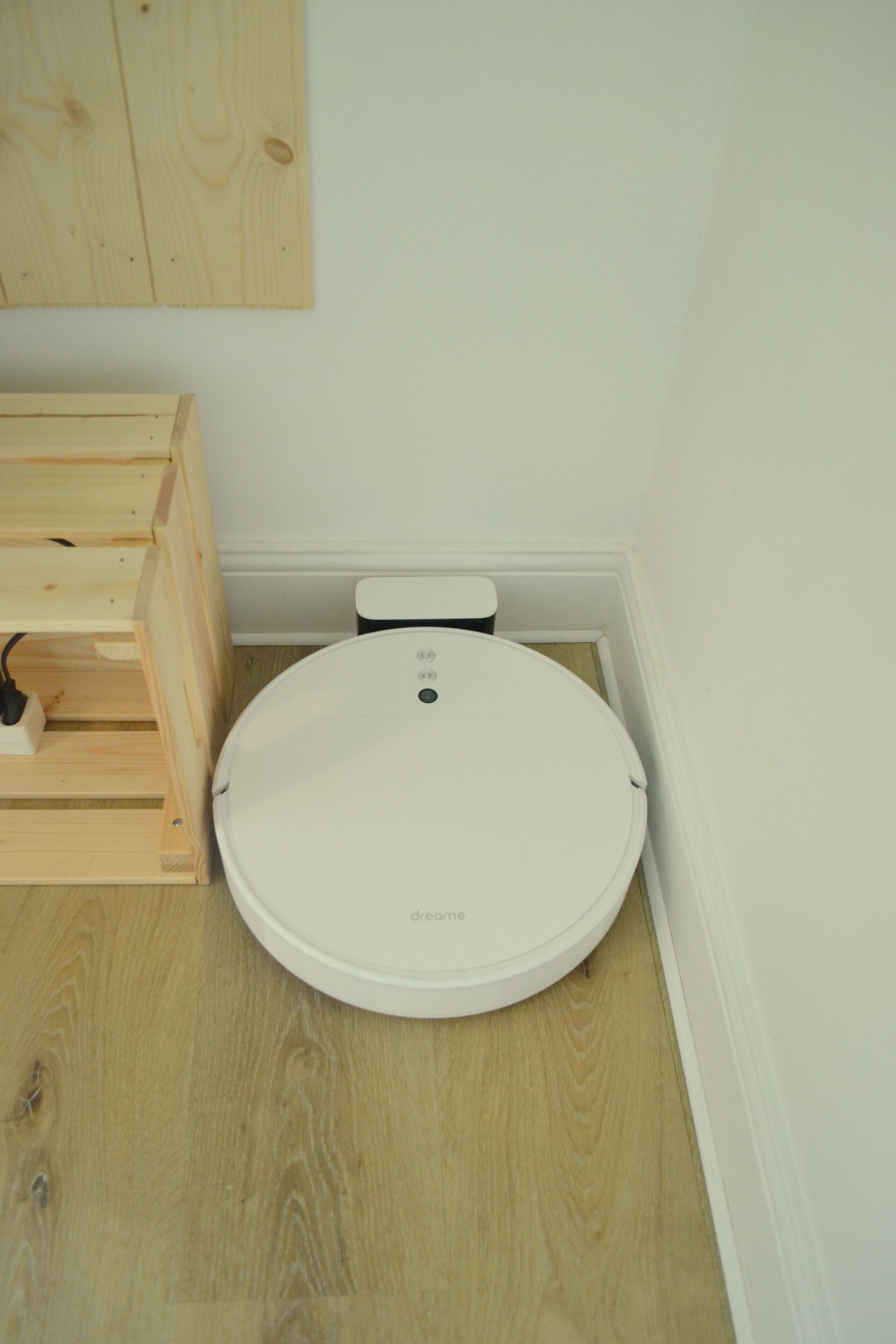 Un dispositivo blanco y circular, compacto, sobre un suelo de madera, probablemente un inodoro inteligente, con un panel de control y un cable de alimentación.