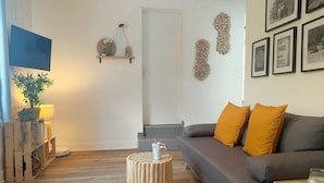 Un salon avec un canapé gris, des coussins de couleur moutarde, une table basse ronde en bois et une petite plante posée sur un support en bois.