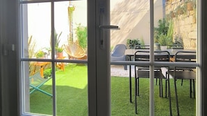 Une vue à travers une porte dotée d’un panneau de verre, révélant un patio avec une table et des chaises, une petite pelouse et un bâtiment avec un mur en pierre et des plantes.