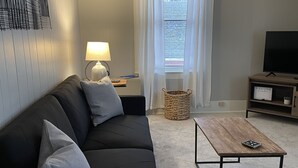 Una sala de estar moderna con un sofá negro, una mesa de centro de madera, una lámpara y un televisor sobre un mueble de madera.