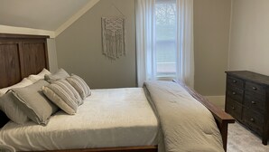 Un dormitorio con cama, cabecera de madera, mesita de noche, cómoda y ventana con persianas.