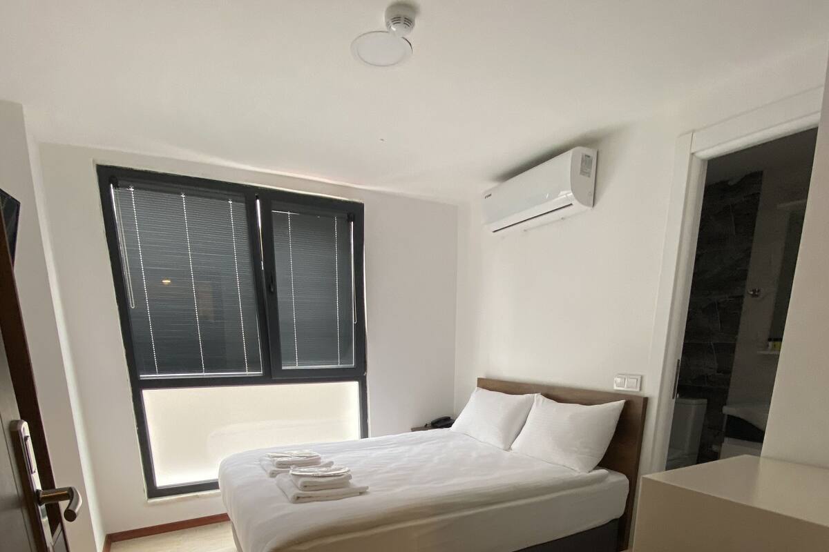 A bedroom with a bed, a window with blinds, a bedside table, and a small desk.