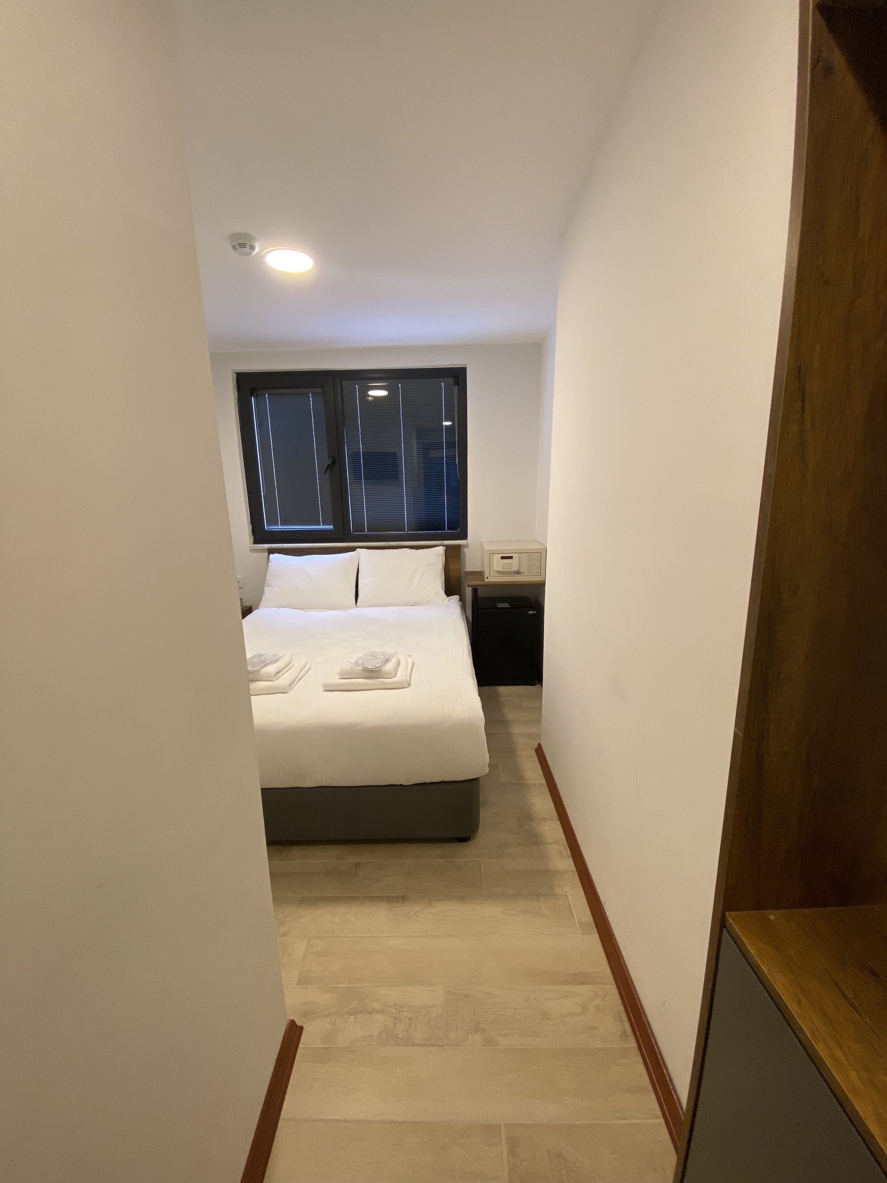 Un pasillo estrecho que conduce a un dormitorio con una cama, una mesita de noche y una ventana con persianas.