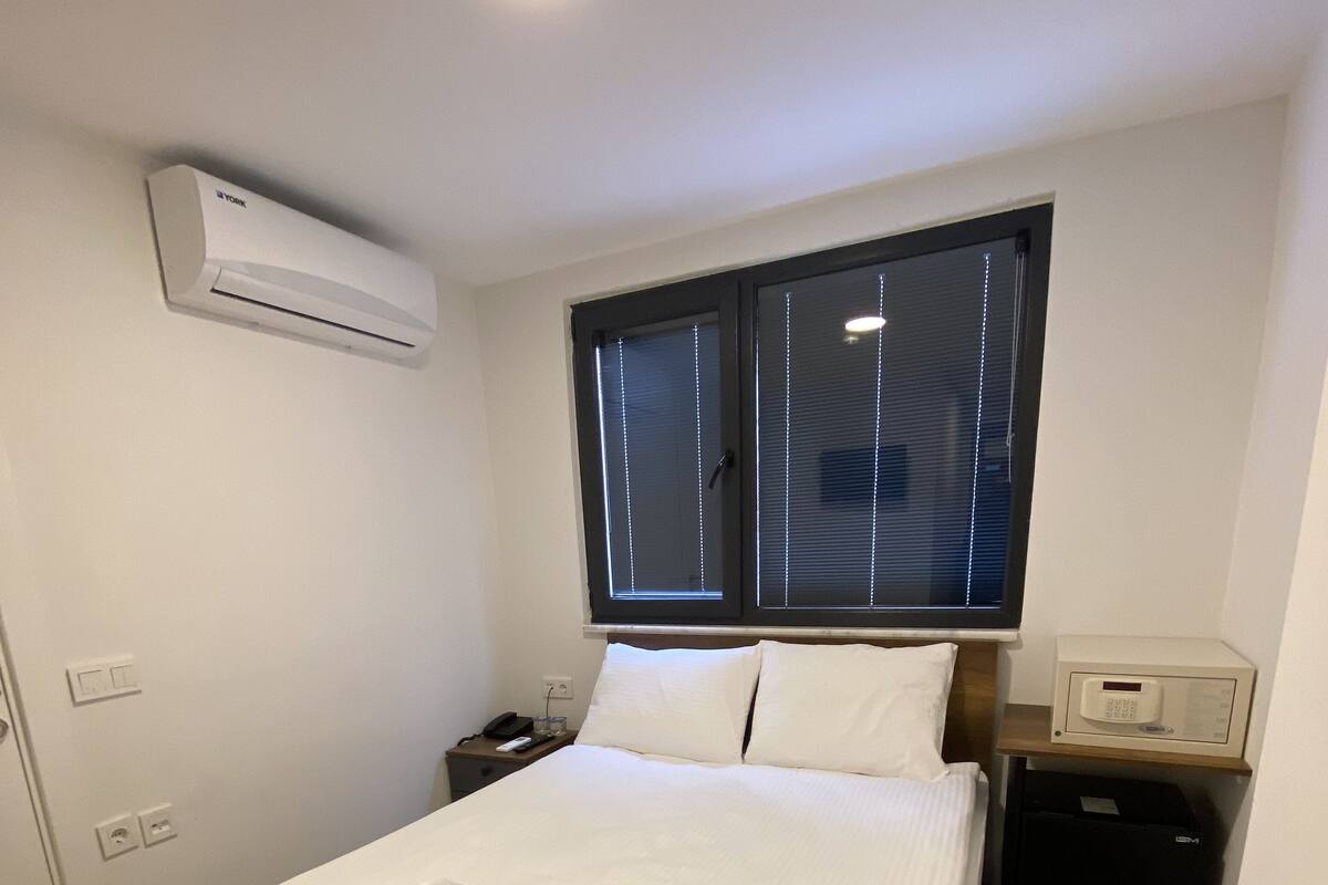 A hotel room with a bed, a nightstand, an air conditioning unit, a window with blinds, and a bedside table with a lamp.