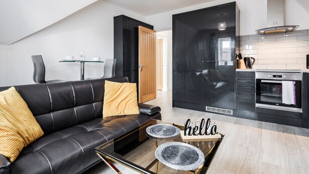 A modern living room with a black leather sofa, a glass coffee table, and a kitchen area with black cabinets and stainless steel appliances. - Comfortable 2BR Apartment in Bedford (Bedford)