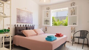 Un dormitorio con cama, una ventana que da a vistas de vegetación, una silla y un estante con toallas.