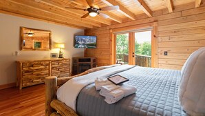 A wooden cabin bedroom with a bed, a ceiling fan, a TV, and a view of the outdoors. - Authentic Log Cabin in the Mountains (Seven Devils)