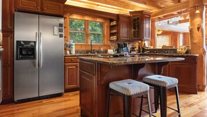 A kitchen with wooden cabinets, a stainless steel refrigerator, a large island with bar stools, and a hanging light fixture. - Authentic Log Cabin in the Mountains (Seven Devils)
