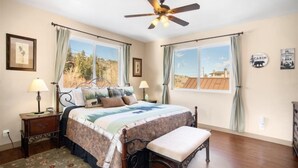 A bedroom with a bed, a ceiling fan, two bedside tables with lamps, a bench, and a view of a cabin through a window. - Hudson Hideaway, your home away from home (June Lake)