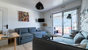 A modern living room with a grey sofa, a coffee table, and a dining table with chairs. A flat-screen TV is mounted on the wall, and there is a large window with a view of the outside.