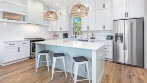 A modern kitchen with white cabinetry, a large island with bar stools, stainless steel appliances, and pendant lighting. - Paradise | Oceanview, Hottub, Shuffleboard, Bikes, Beach, Pool (Port St. Joe)