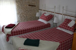 Une chambre avec deux lits, chacun recouvert d’une courtepointe à carreaux rouges et blancs, agrémentée d’oreillers blancs et dotée d’une tête de lit en bois. On y trouve un mur en pierre et une fenêtre avec des rideaux.