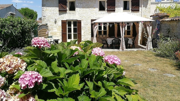 A stone house with a covered patio, a garden with hydrangeas, and a neighboring building with blue shutters. - Beautiful Gite with pool and jacuzzi 6 pers 3 bedrooms (Nantille)