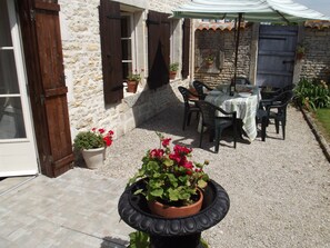 Ein Steinhaus mit einer Terrasse, auf der sich ein Tisch, Stühle, Kübelpflanzen und ein mit Streifen versehener Pergola befinden.