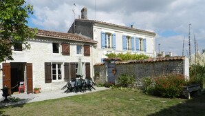 A two-story stone building with a tiled roof, a small garden with a table and chairs, and a stone wall with a window. - Beautiful Gite with pool and jacuzzi 6 pers 3 bedrooms (Nantille)