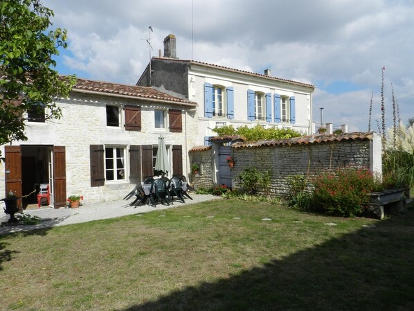 Un bâtiment en pierre de deux étages, avec un toit en tuiles, un petit jardin agrémenté d’une table et de chaises, et un mur en pierre percé d’une fenêtre.