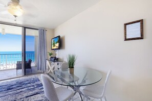 A room with a glass table and chairs, a TV mounted on the wall, and a view of the beach through a sliding glass door. - Beachfront Condo | Restaurant, Pool, Spa & Gym (Marco Island)