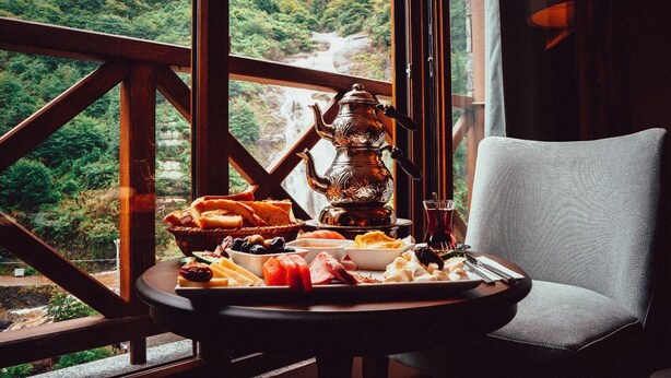 A table with a tea set and various foods, a chair, and a view of a mountainous landscape through large windows. - Grand Selale Hotel (Çayeli)