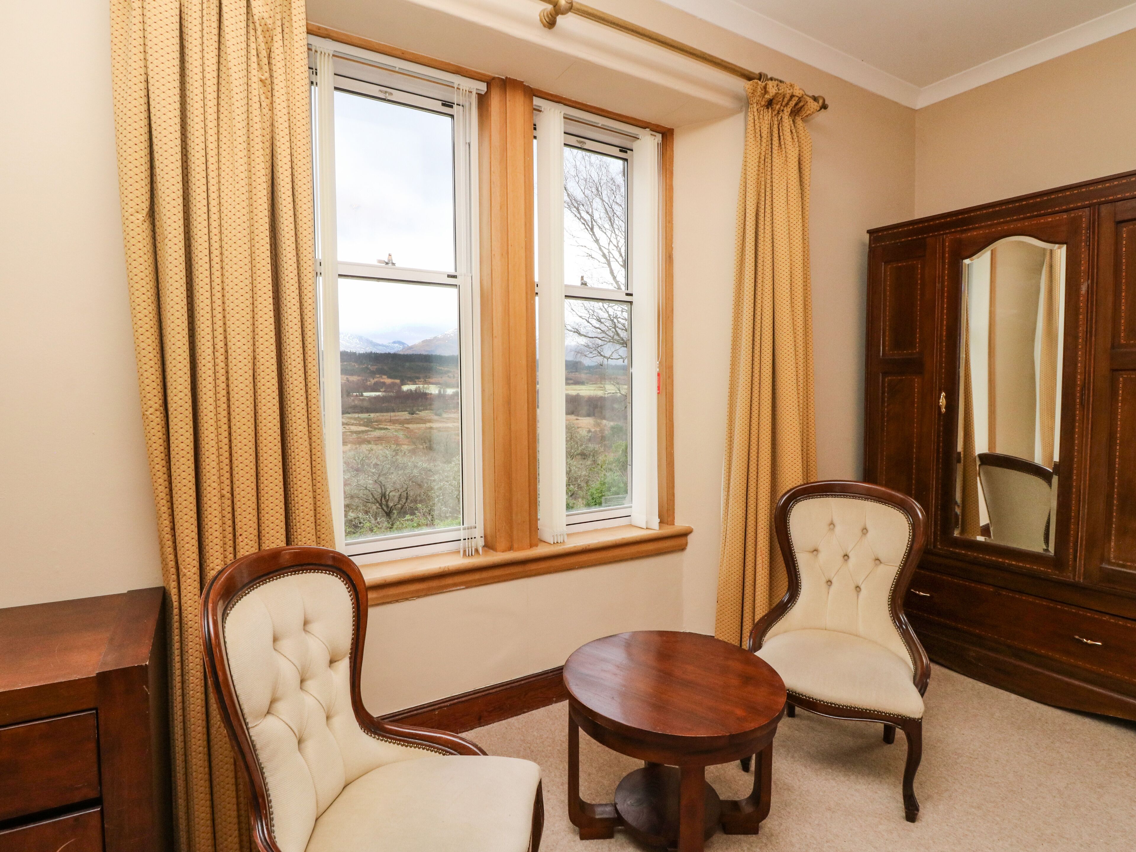 A room with two upholstered chairs, a wooden table, a large mirror, and a window with curtains.