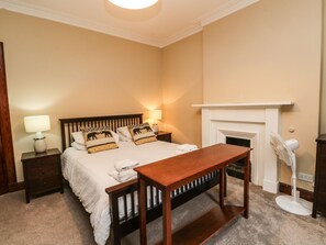 Ein Schlafzimmer mit einem Bett, Nachttischen, einer Kommode, einer hölzernen Bank, einem Standventilator und einem Kamin.