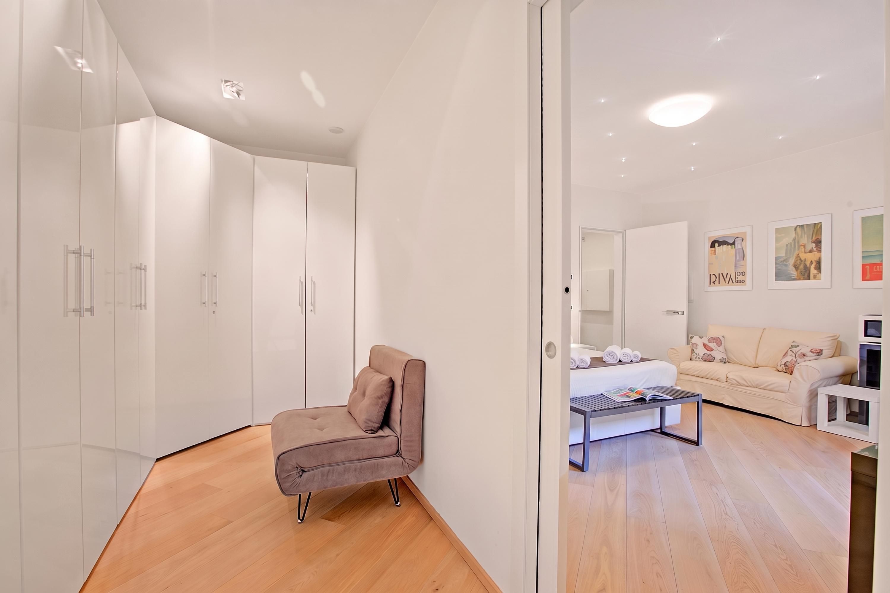 Een moderne slaapkamer met een bed, een bank en een stoel. Er staan witte kasten en er hangt een spiegel.