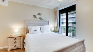 A bedroom with a wooden headboard, a bed with white bedding, a nightstand with a lamp, and a window with a view of a building. - White Caps 201 by Liquid Life (Orange Beach)