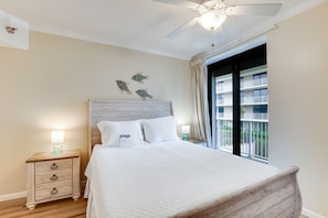 Un dormitorio con cabecera de madera, cama con ropa blanca, mesita de noche con lámpara y ventana con vista a un edificio.