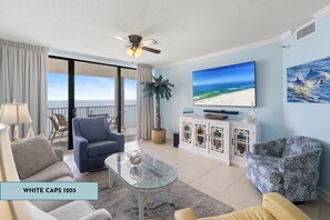 A spacious living room with a glass coffee table, a blue armchair, a beige sofa, and a patterned armchair. A large flat-screen TV is mounted on the wall, and a ceiling fan is visible. - White Caps 1203 by Liquid Life (Orange Beach)