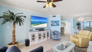 Una sala de estar con ventilador de techo, televisor grande, palmera y comedor con sillas.