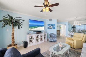 Un salon doté d’un ventilateur de plafond, d’un grand téléviseur, d’un palmier et d’un coin repas avec des chaises.