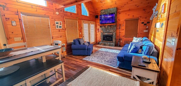 L’intérieur chaleureux d’un chalet avec une cheminée en pierre, deux canapés, une table à manger et un téléviseur fixé au-dessus de la cheminée.