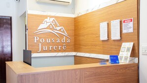 A reception desk with a wooden finish, a logo with a house and waves, and a sign with 'AVISO' and a list of services. - Pousada Jurerê (Florianópolis)